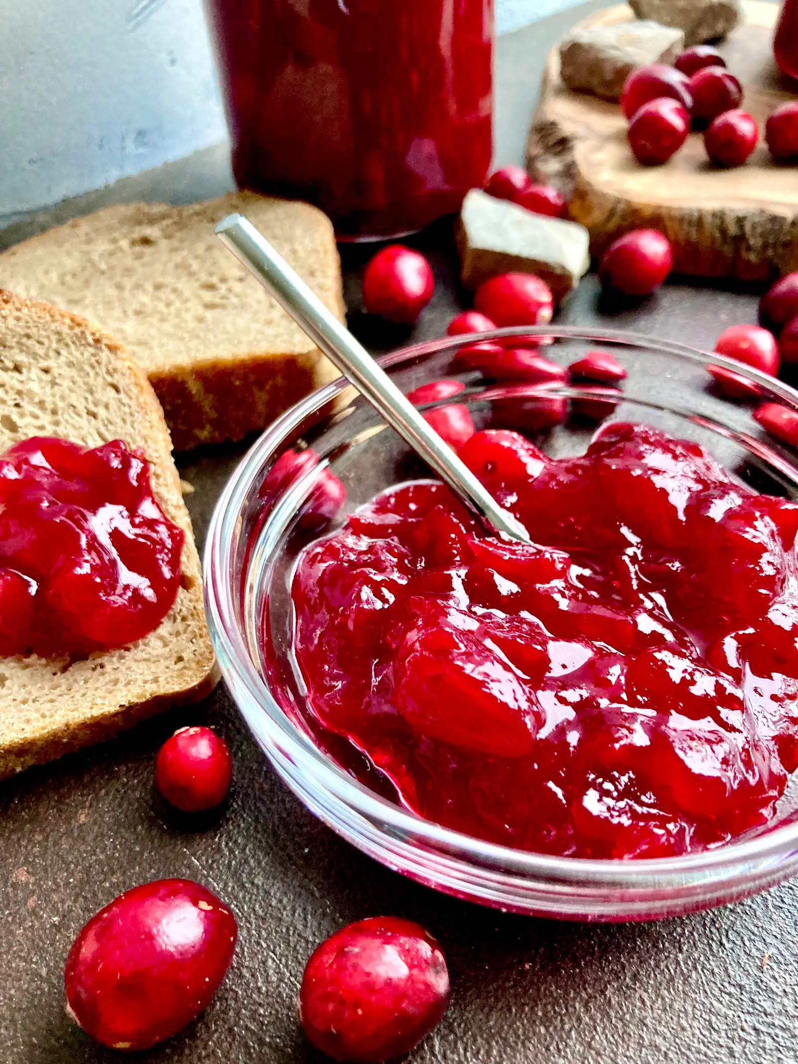 Marmelade mit Cranberries in kleiner Glasschale mit silbernen Löffel darin dahinter aufgeschnittenes Brot und Cranberries